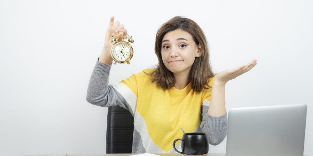 Photo of female office worker sitting at the desk and holding alarm clock . High quality photo