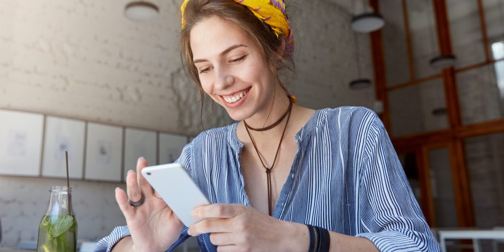 Happy student girl with charming smile reading positive news or browsing newsfeed via social networks using mobile phone, having small break while doing homework, sitting at cafe and drinking lemonade